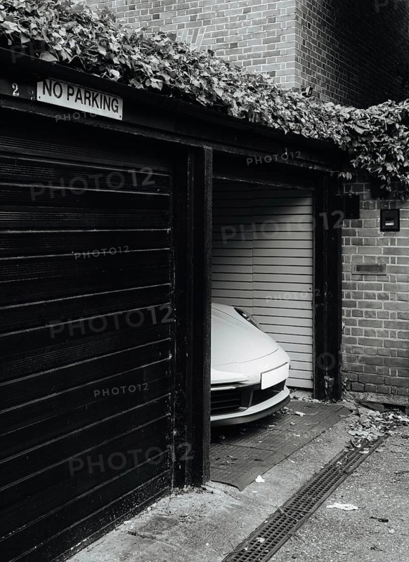 A sleek sports car is discreetly parked in a garage with a No Parking sign. The urban London setting features brick walls and foliage for contrast and intrigue