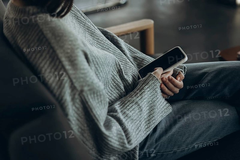 A person wearing a comfy sweater and jeans relaxes on a sofa, holding a smartphone. The setting exudes a cozy, casual home environment, perfect for unwinding