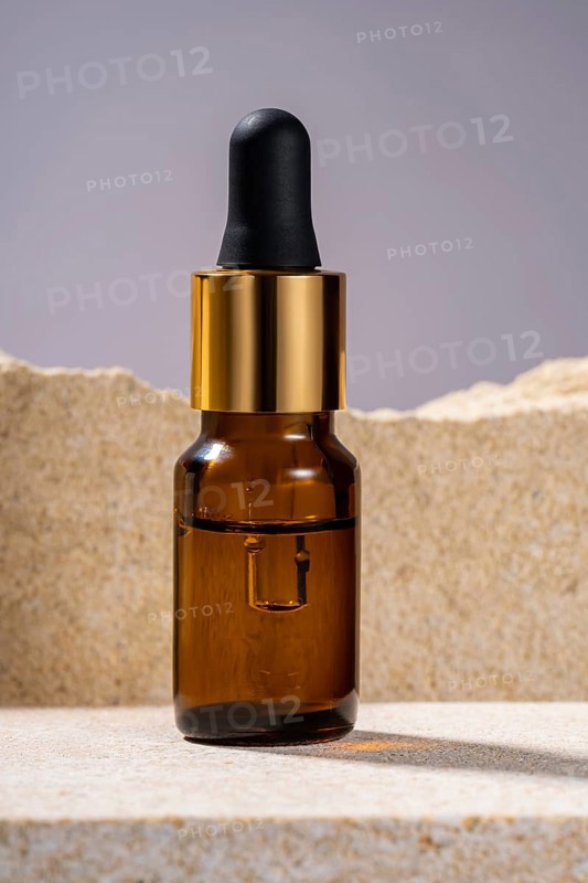 A stylish amber glass serum bottle with a black dropper and golden details, showcased on a textured stone podium, ideal for luxury skincare branding