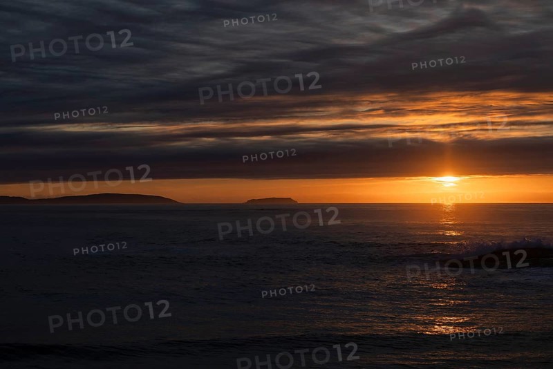 A serene sunset over the ocean, with the sun casting radiant orange and yellow hues across the sky. Silhouetted islands in the distance add to the tranquil beauty, with calm waters reflecting the vibrant colors amidst the dark, cloudy sky