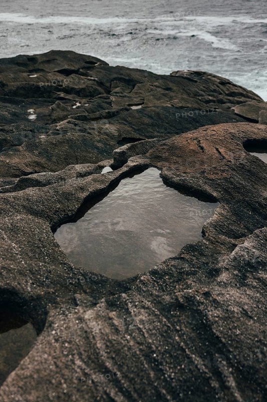 A serene coastal scene features rugged rock formations with shallow pools of water collected within the crevices. The ocean waves create a tranquil, yet dynamic backdrop, highlighting the natural beauty of the shoreline