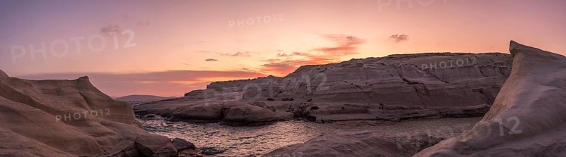 Sweeping panoramic view of Sarakiniko beach on Milos Island, Greece, under a vibrant sunset sky. The unique white volcanic rock formations gracefully surround a tranquil sea