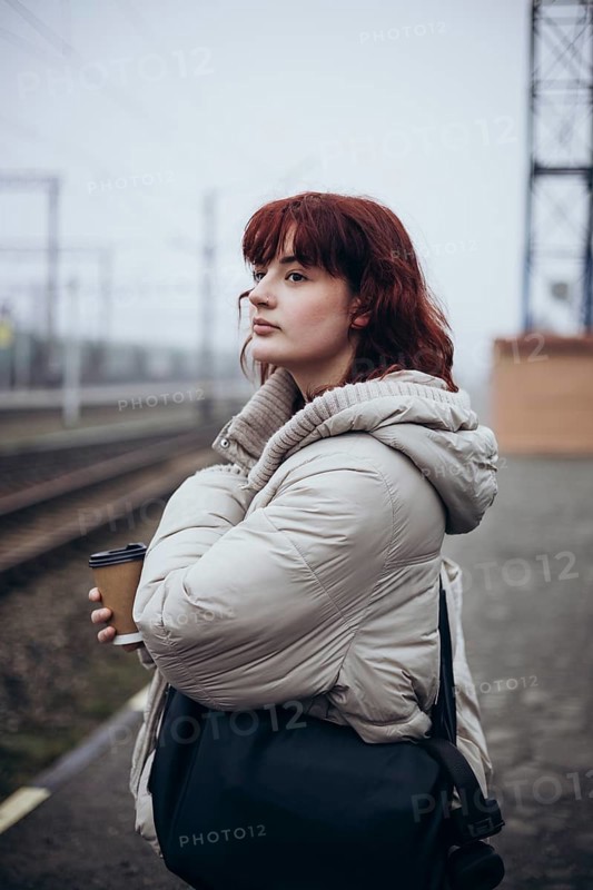 A young woman in a white coat stands at a train station, holding a coffee cup. The scene is foggy, creating a serene atmosphere, perfect for depicting travel or waiting themes