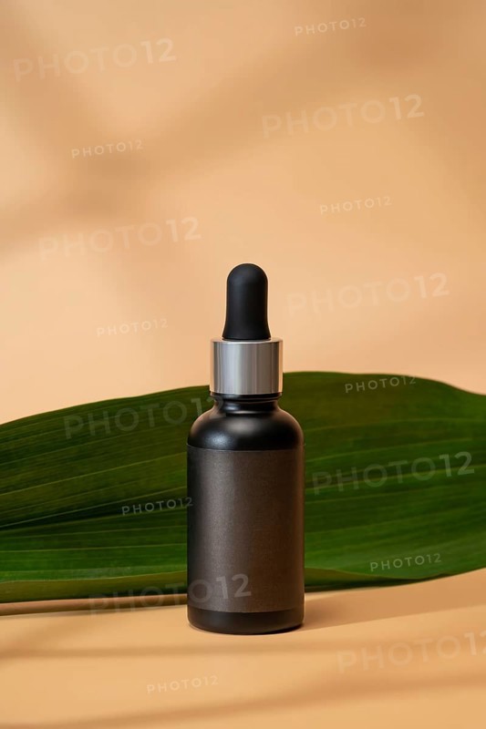 A sleek cosmetic bottle with a dropper cap sits elegantly on a soft beige background. A green leaf adds a natural element, enhancing the minimalist and tranquil design
