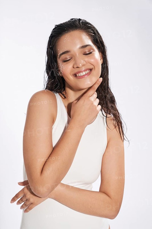 Studio portrait of a joyful female model with wet hair and skin, her eyes gently closed, wearing a white bodysuit, embodying freshness and beauty