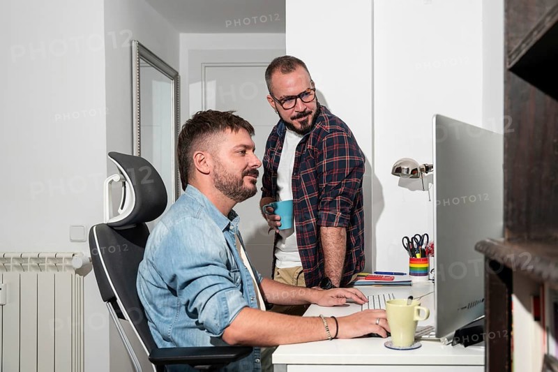 Two gay men collaborate in a cozy home office, mixing work with casual interaction. One sits at a desk while the other stands beside him, both focused and in good spirits