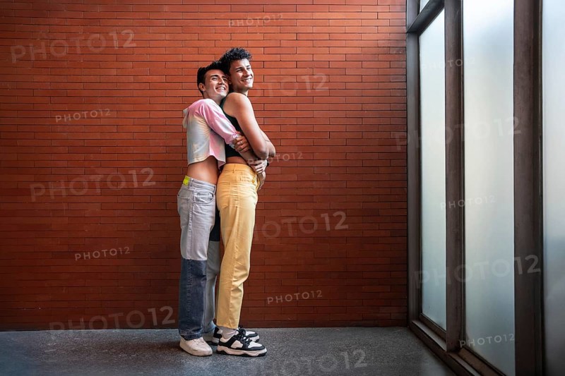 A joyful gay couple embraces lovingly against a brick wall. Their happy expressions and casual attire convey warmth, affection, and a sense of togetherness in natural light