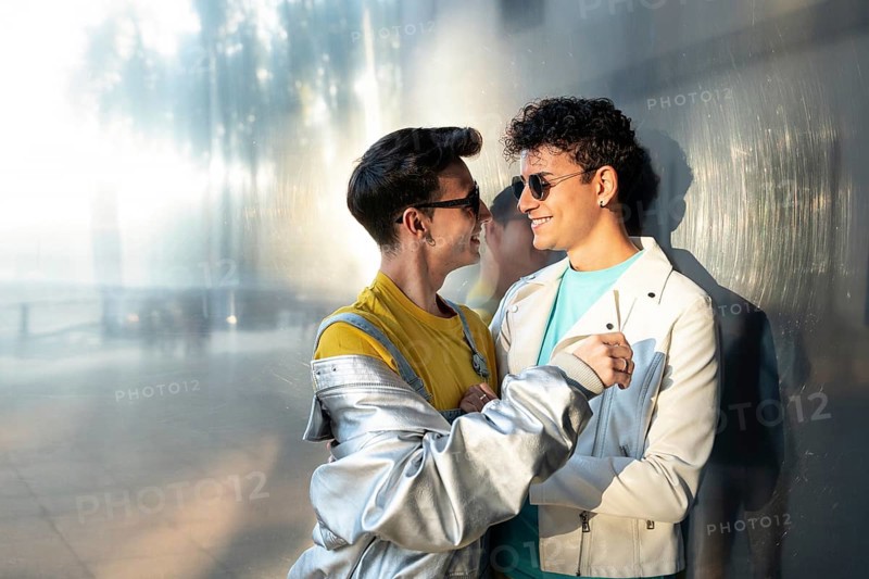 A joyful gay couple, wearing casual clothes and sunglasses, share a tender moment while smiling at each other. The scene is warm and vibrant, capturing love and connection