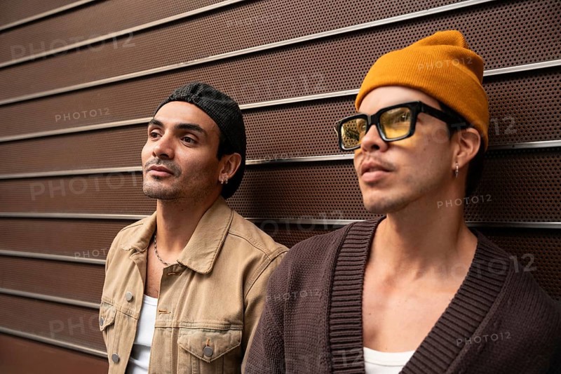 A colombian gay couple stands side by side against a modern metal wall. Both are dressed in casual urban attire, with one wearing a beanie and glasses, the other a cap