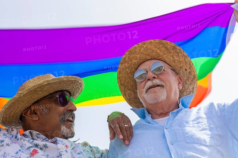 From below joyful gay senior couple, comprising two men, is captured outdoors, sharing a happy moment under a colorful rainbow flag. They are both wearing hats and sunglasses, with one man smiling at the other