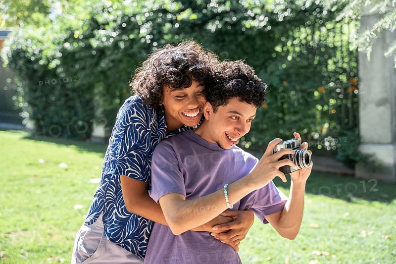 A happy multiethnic LGBTQ+ couple embraces while taking photos in a sunny park. Their joyful expressions reflect love and togetherness, capturing a moment of shared happiness
