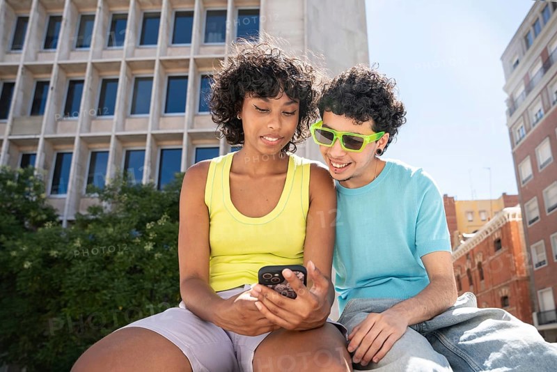 A multiethnic LGBTQ+ couple enjoys a sunny day in the city, looking at a smartphone together. The backdrop features modern architecture and urban vibes, capturing a lively moment
