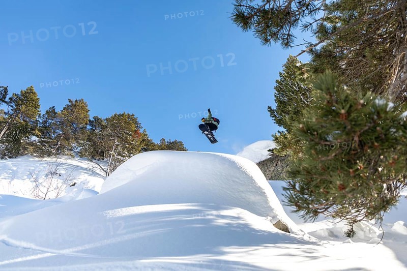 A snowboarder soars through the clear blue sky, performing an aerial trick above a snowy mountain landscape surrounded by evergreen trees. The scene captures the thrill of winter sports