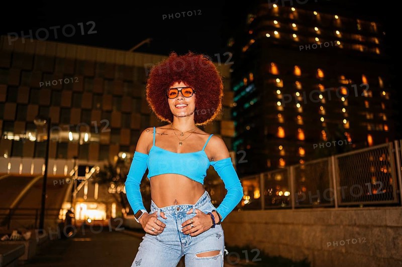African descendent woman with striking afro hair and sunglasses poses confidently outdoors at night with urban buildings illuminated in the background