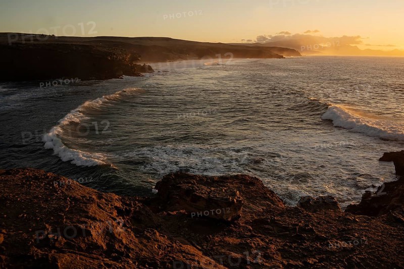 Dramatic view of powerful waves hitting rugged rocks with a sunset backdrop at La Pared, Fuerteventura. The warm golden light enhances the scenic beauty of the coastal landscape
