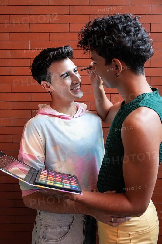 A couple shares a tender moment as one applies makeup to the other with a colorful palette, set against a brick wall, expressing love and creativity