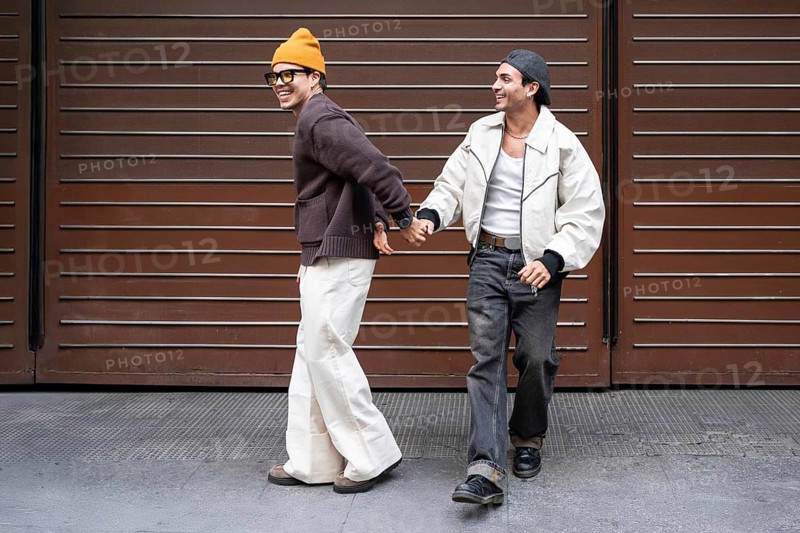 A joyful couple holds hands while walking in an urban setting. They are smiling, showcasing happiness and connection. One wears a beanie, the other a cap, exuding casual style