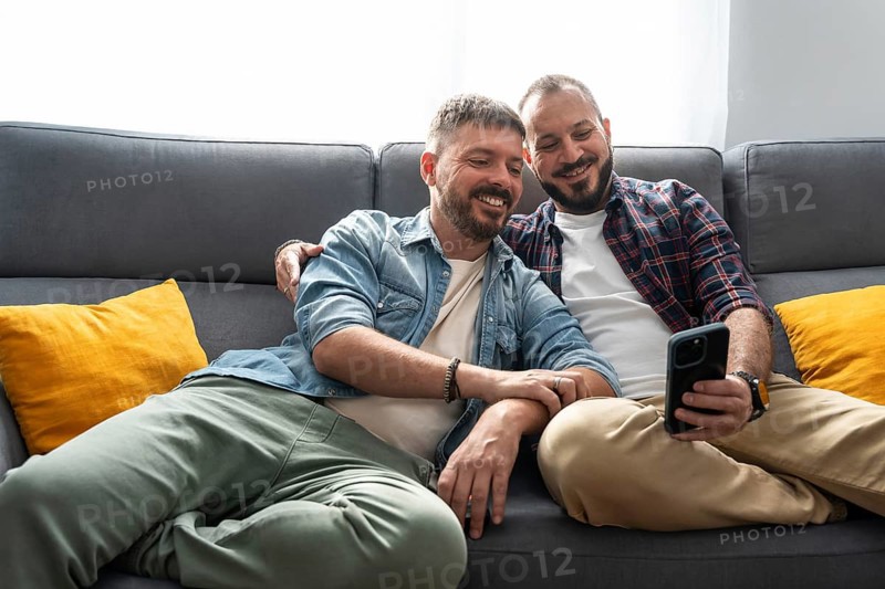 A joyful gay couple sits comfortably on the sofa, smiling as they look at a smartphone together in a cozy living room