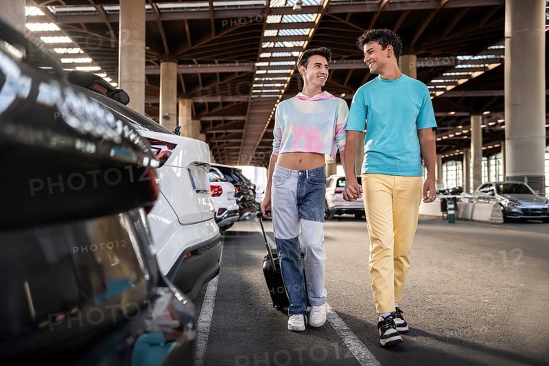 A gay couple walks hand in hand through a sunny parking area filled with cars. Both are smiling and enjoying a moment of togetherness, symbolizing love and connection