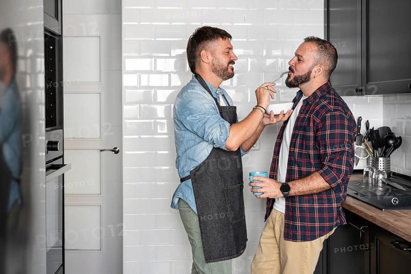 A joyful gay couple shares a moment in the kitchen, one feeding the other with a fork while smiling. They embody love and domestic bliss in a modern cooking space