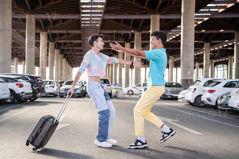 A cheerful gay couple reunites in a spacious parking lot, one carrying a suitcase. They are embracing, expressing happiness and love in a lively urban setting