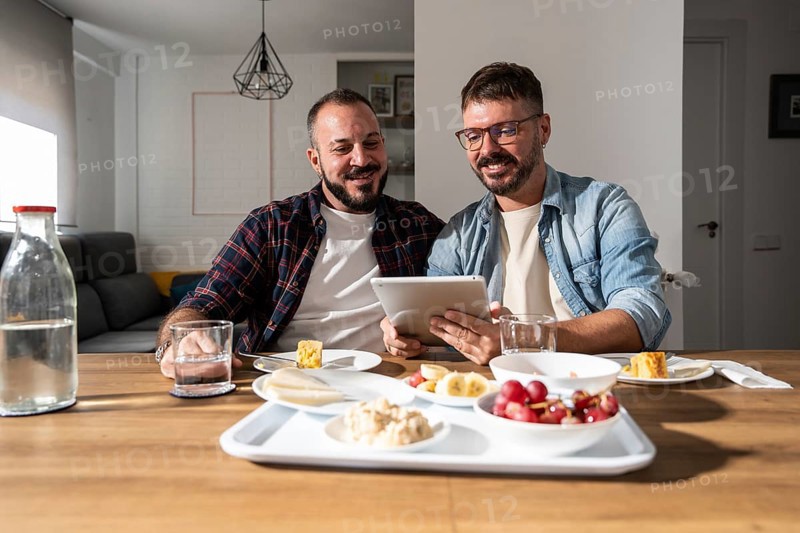 A gay couple enjoys breakfast together at a table, sharing a tablet and smiling. Fresh fruit, water, and pastries are on the table, with soft light illuminating the cozy room