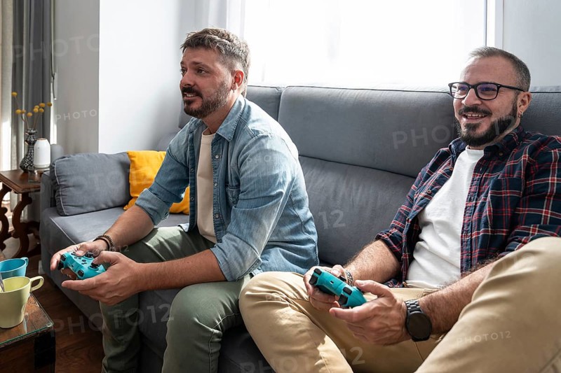 A gay couple is sitting on a sofa, joyfully playing video games together in a bright and cozy living room setting