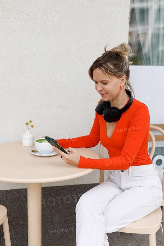 A woman sits at an outdoor cafe in Warsaw, using her smartphone and wearing headphones She enjoys a cup of matcha while engaging with her gadgets in a relaxed environment