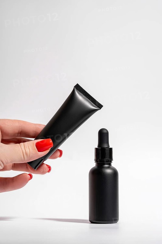 Cropped unrecognizable female hand with red manicure holding a matte black tube and dropper bottle isolated on a white background