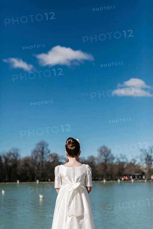 A young girl in a white First Communion dress stands by a tranquil lake, gazing at the serene view. The clear blue sky and peaceful water create a sense of calm