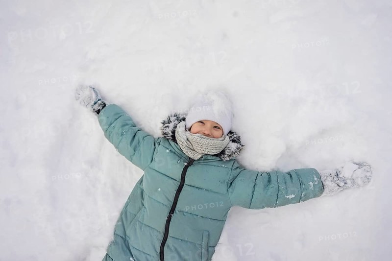 A child joyfully lies in the snow, wearing a green coat, white hat, and scarf. The scene captures the essence of winter fun and the coziness of cold weather attire
