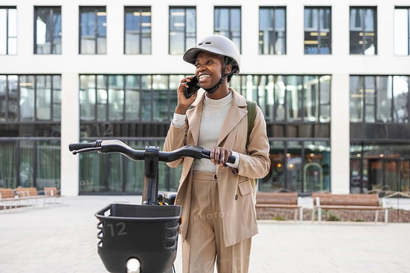 Smiling black woman in sleek beige suit and helmet, talking on a smartphone while holding a bike in a modern urban environment Confident and stylish city life