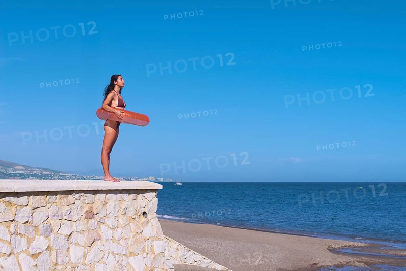 Side view of a young woman inside a float while standing atop a stone wall at the beach, gazing over a calm sea against a clear blue sky