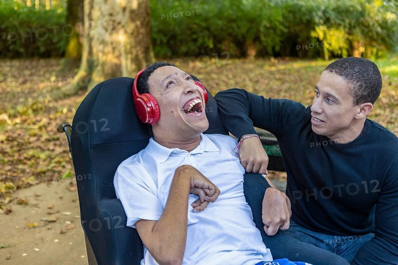 Spanish Cameroonian siblings share a joyful outdoor moment at a park. One sibling, wearing headphones, has cerebral palsy, highlighting the family's Mixed-race bond