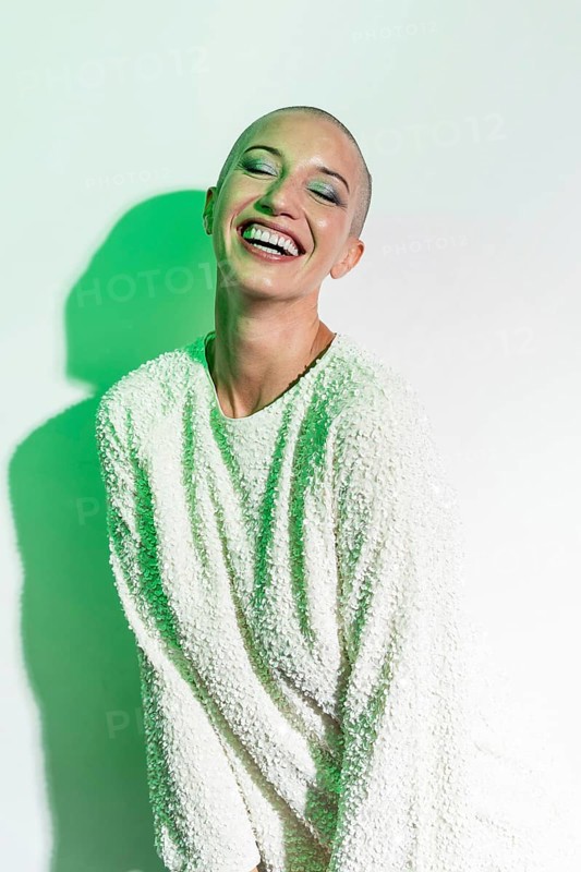 A cheerful middle-aged woman with a shaved head smiles brightly. She wears a textured white sweater, standing against a light background with green lighting