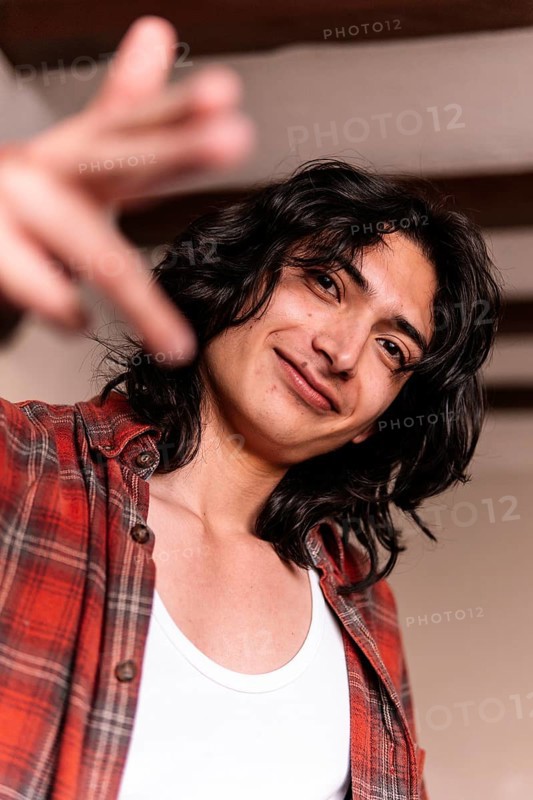 A smiling young man in a white tank top and plaid shirt extends his hand towards the camera, giving a friendly and inviting gesture in a warmly lit indoor setting