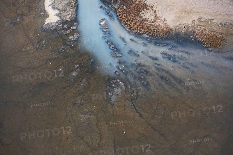 A detailed aerial shot of a lagoon in Toledo, Spain, displaying muddy textures and branching water channels. The natural design of sediment and water flow creates an intricate, organic pattern