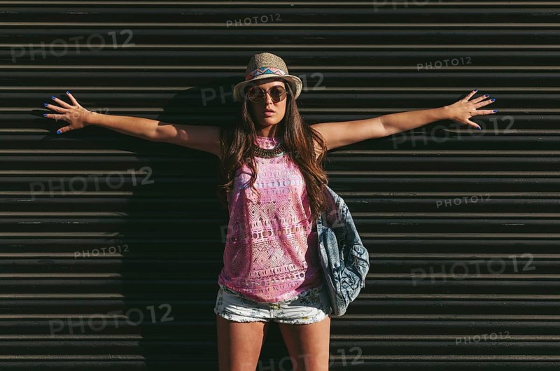 A fashionable woman poses confidently against a dark background, wearing a pink sleeveless top, shorts, a hat, and sunglasses, exuding summer style and confidence