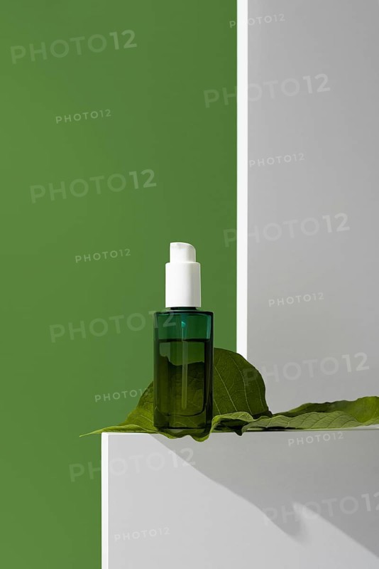 Close-up view of a green glass essential oil bottle placed on a white stand with a large green leaf, set against a vibrant green background. The natural setting emphasizes the product's use in skincare and wellness
