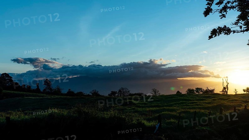 Capture the serene beauty of sunset over lush green fields under a vibrant sky lit by the descending sun, with misty mountains in the distance