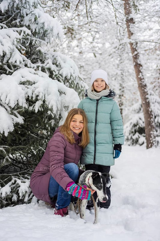 A joyful family poses with a dog in a snowy forest, surrounded by snow-covered trees. The bright winter attire contrasts with the serene white landscape