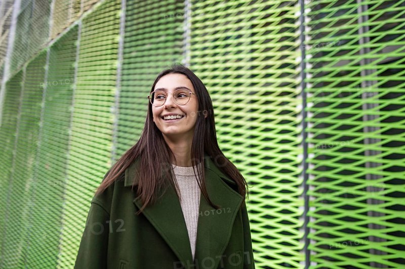 A woman in a stylish green coat smiles joyfully while standing against a vibrant, abstract green background Her glasses add sophistication to the modern urban setting