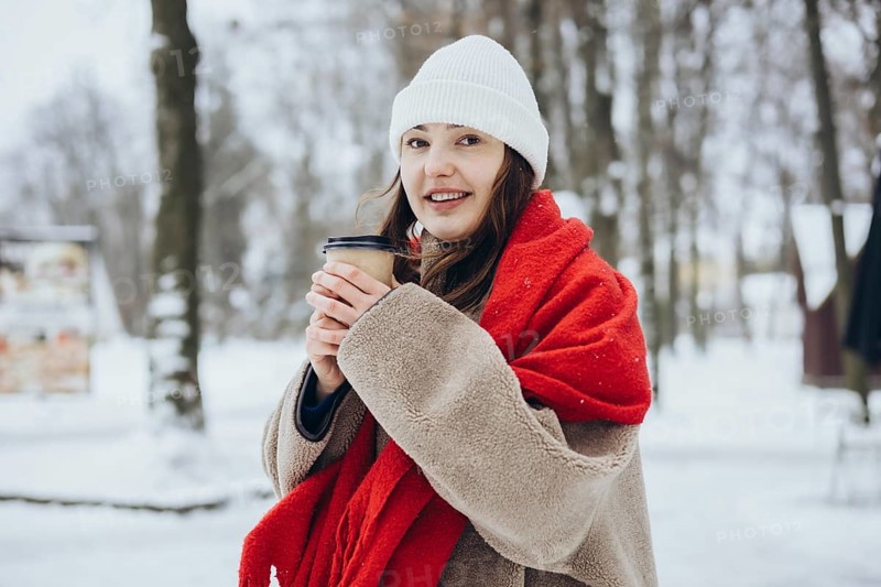 A young woman in winter clothing holds a coffee cup while wrapped in a cozy, red scarf, in a snowy landscape