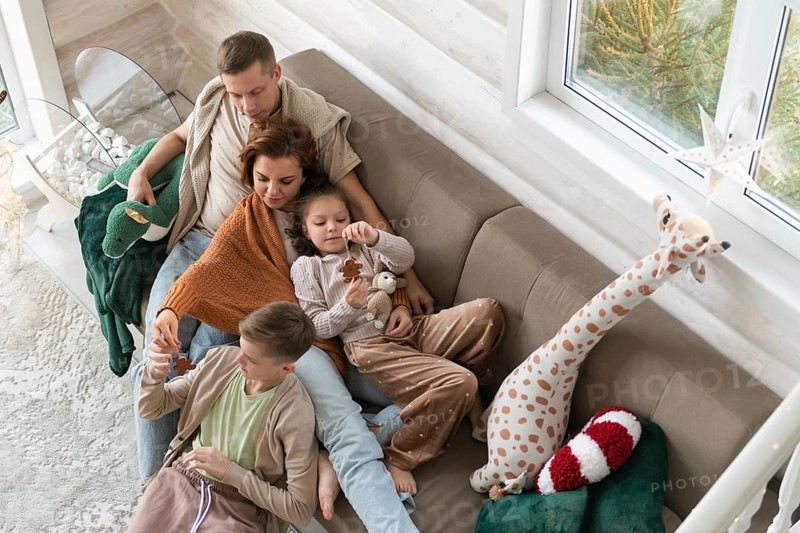 Top view of a festive setting with a winter view and holiday deco. A joyful family celebrates Christmas together on a couch. A couple relax with two kids, an older boy and a younger girl. Both children are engrossed with toys