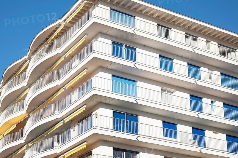 A modern apartment building features multiple curved balconies, white facade, and glass railings, set against a clear blue sky, showcasing urban architecture