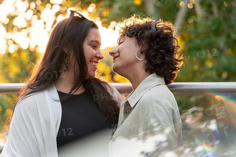 A lesbian couple shares a tender moment, smiling warmly at each other in the soft glow of sunset, symbolizing love and connection against a backdrop of vibrant greenery