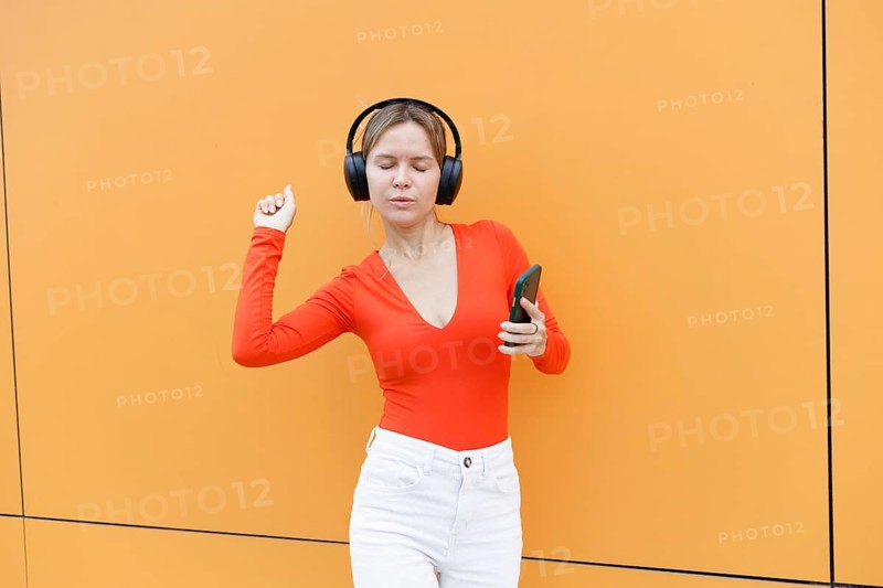 Woman in a red top using a smartphone and wearing headphones, standing against an orange wall in Warsaw She appears to be enjoying music or a podcast