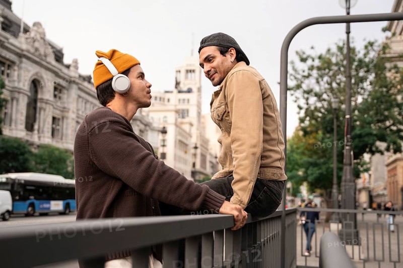 A Colombian couple enjoys a moment together in Madrid city, one listening to music with headphones, the other sitting on a railing with vibrant expressions