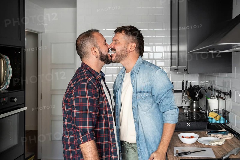 A joyful gay couple in a kitchen, playfully sharing cream from a dessert, showcasing love and happiness in a cozy home setting