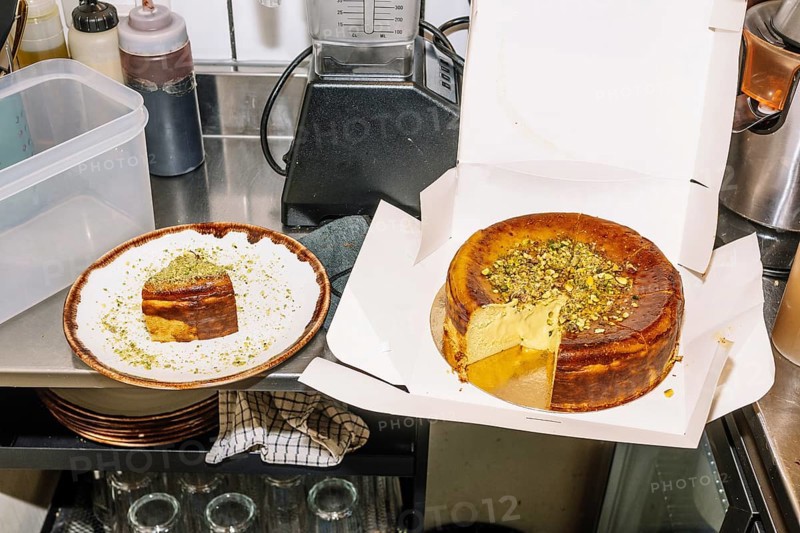 A rich cheesecake topped with crushed pistachios sits in an open box, with a slice served on a plate nearby. The scene is set in a coffee shop kitchen with baking tools and ingredients